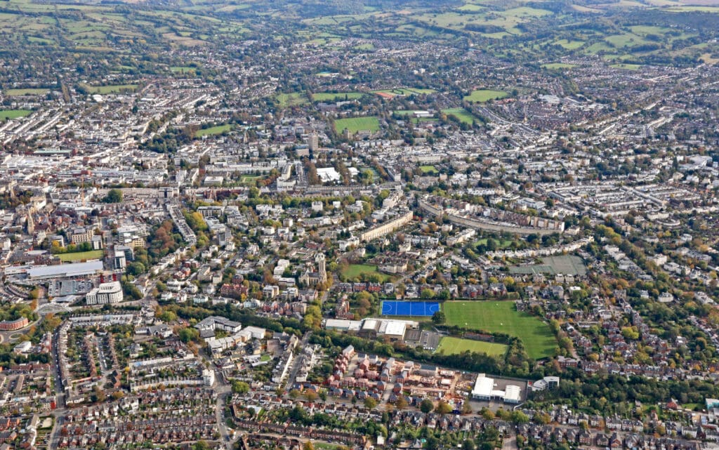 Aerial view of a large town with densely packed houses, green parks, sports fields, and tree-lined streets, surrounded by rolling hills and countryside in the distance.