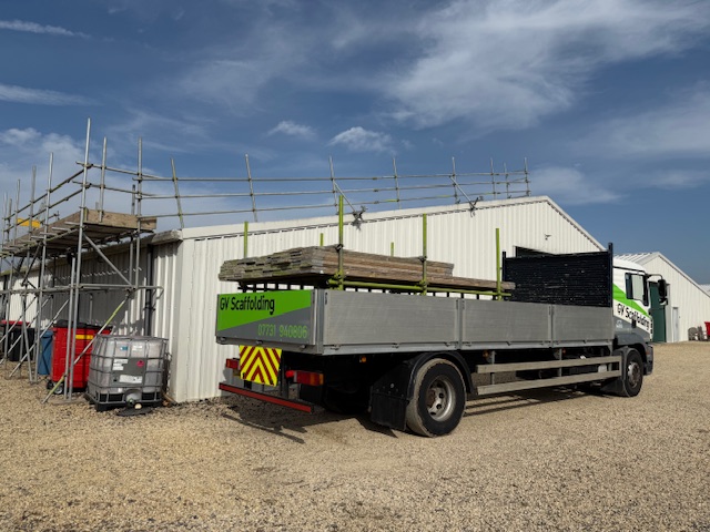A flatbed truck labeled "GJ Scaffolding" is parked on gravel beside a white industrial building. Scaffolding and construction materials are stacked nearby under a clear blue sky.