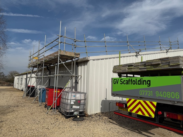A metal building with scaffolding erected along its side. A truck with “GV Scaffolding” and a phone number printed on it is parked nearby. Stacks of materials and large water containers are on the ground. The sky is partly cloudy.