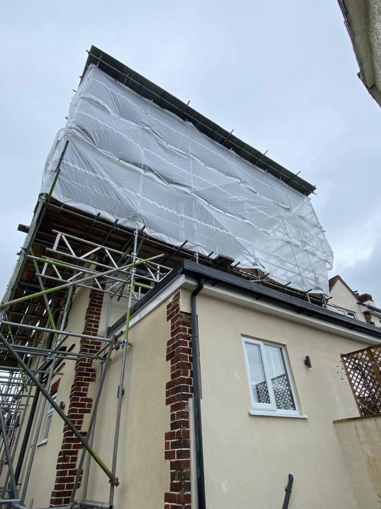 Scaffolding covered with white plastic sheeting surrounds the upper part of a house, indicating ongoing construction or renovation work. The sky is overcast.
