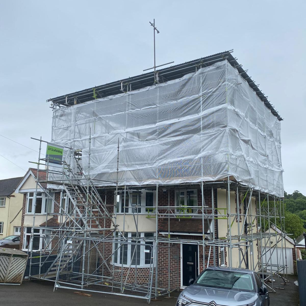 A two-story brick house is covered in scaffolding and wrapped in white sheeting for construction or renovation work. Stairs lead to the upper level, and a car is parked in front of the house.