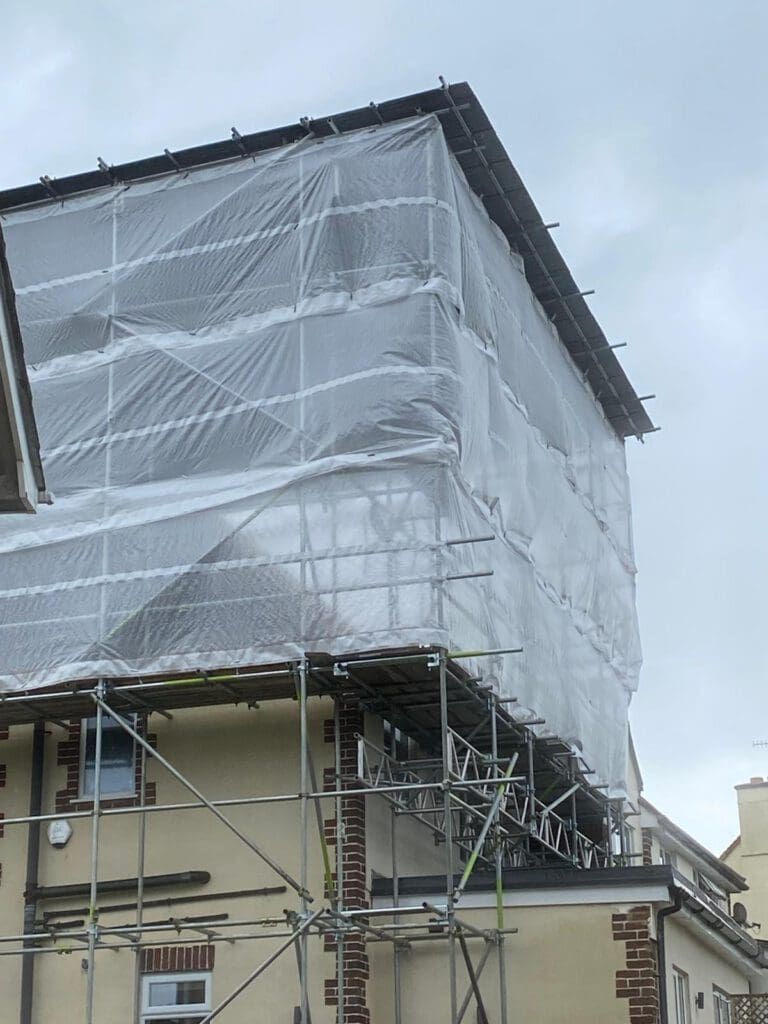 A building covered in scaffolding and wrapped in white plastic sheeting, likely for construction or renovation work, against a cloudy sky.