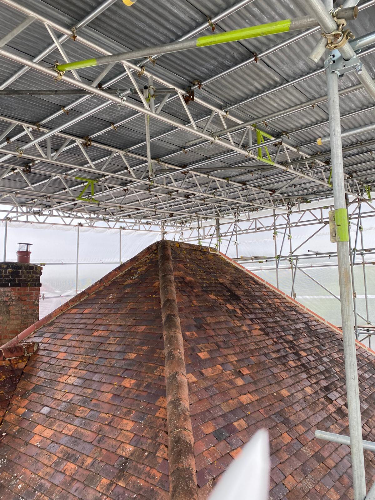 A view of a pitched, weathered tiled roof under a large scaffolding structure covered with corrugated metal sheets, providing shelter and support for construction or maintenance work.