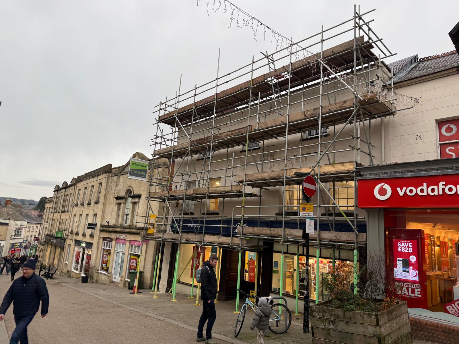 A street scene showing a building covered in scaffolding for construction, with pedestrians walking by. Next door is a Vodafone store with a red sign and sale posters in the window. A bike is parked nearby.