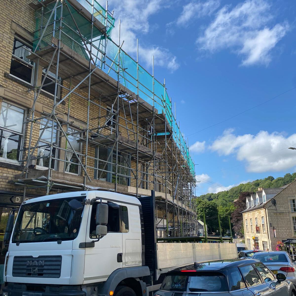 A white truck is parked next to a building covered in scaffolding. Several cars are parked on the street. The sky is mostly clear with a few clouds, and trees are visible in the background.