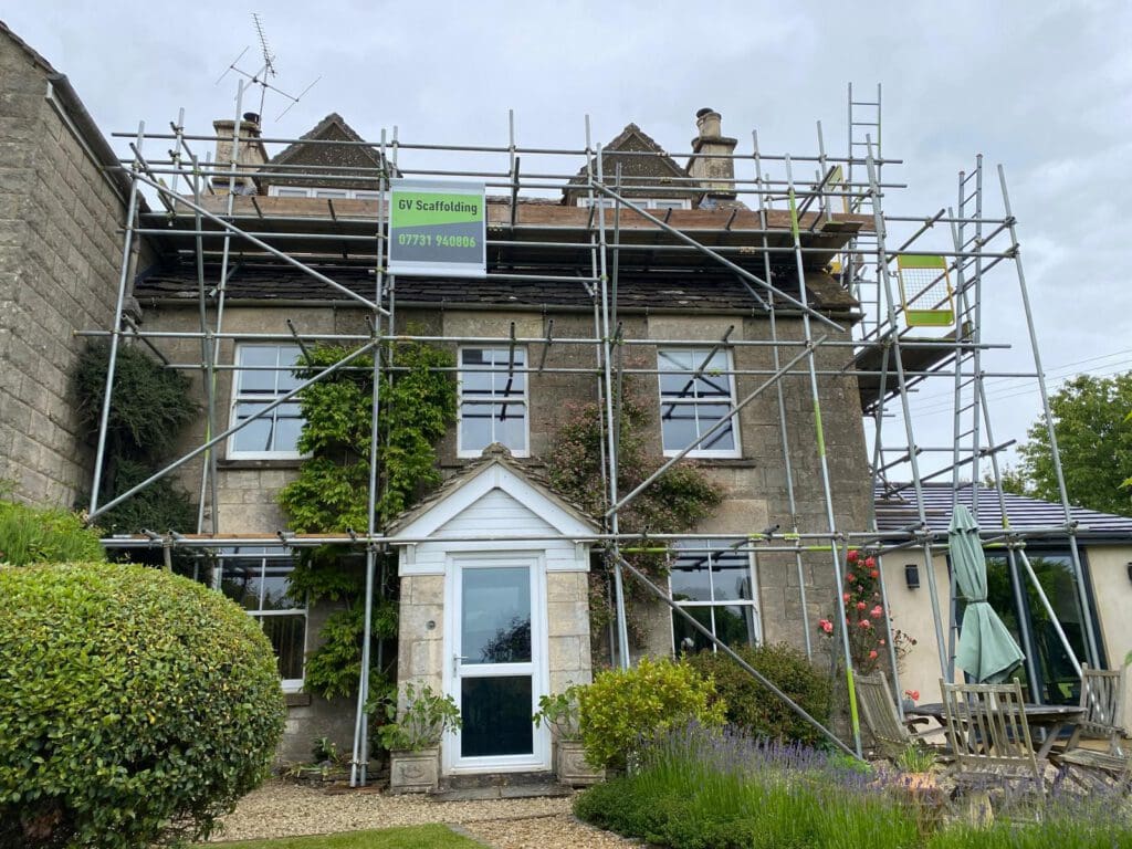 A stone house with a white door and several windows is surrounded by metal scaffolding for construction, with a green GV Scaffolding sign on top. There are bushes, flowers, and outdoor furniture in the garden.