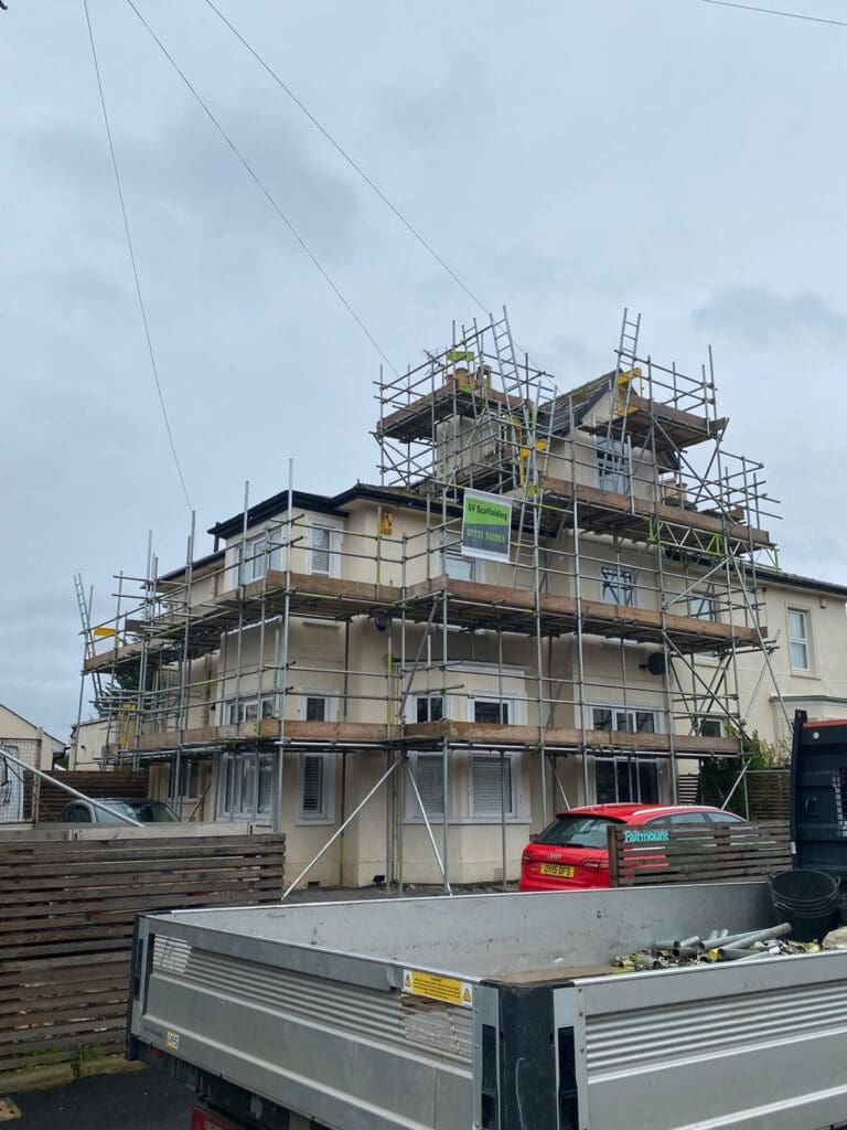 A multi-story building under renovation with scaffolding along its exterior. A red car and a flatbed truck are parked in front. The sky is overcast and stacks of wooden planks are visible near the building.