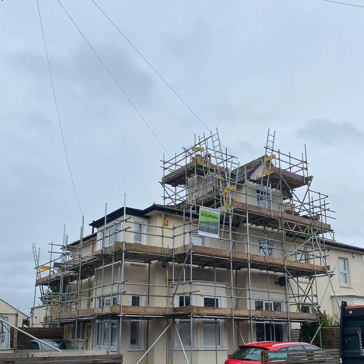 A two-story cream-colored house is surrounded by scaffolding, indicating construction or renovation work. The sky is overcast, and a red car is parked in front of the building.