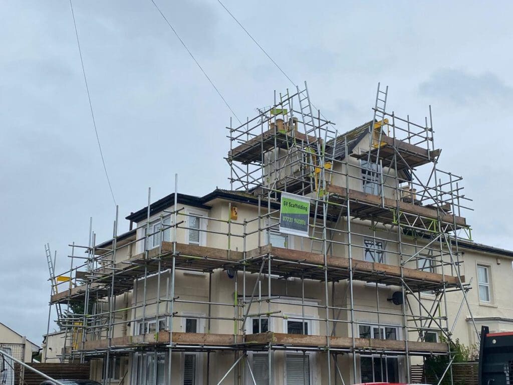A cream-colored multi-story house is covered in scaffolding, likely for construction or renovation work. The sky is overcast, and a sign with contact information is attached to the scaffolding.