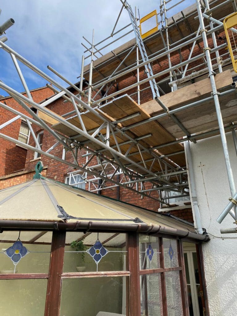 Scaffolding is set up around a brick building above a small glass-roofed sunroom with decorative blue and yellow window panes. The sky is partly cloudy.