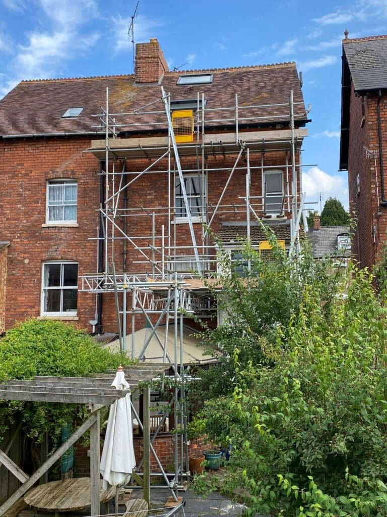A red-brick house with scaffolding covering the back, reaching up to the roof. There is a garden with green bushes and a wooden pergola in the foreground under a partly cloudy sky.