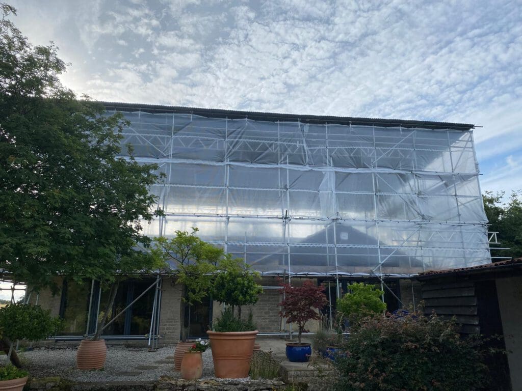 A large building covered in transparent scaffolding sheeting, with trees, potted plants, and a cloudy sky in the background. The structure appears to be under construction or renovation.