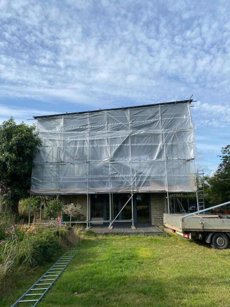 A house covered in scaffolding and transparent plastic sheeting, likely under construction or renovation, with a ladder lying on the grass and a trailer parked nearby. Trees and grass surround the area under a partly cloudy sky.