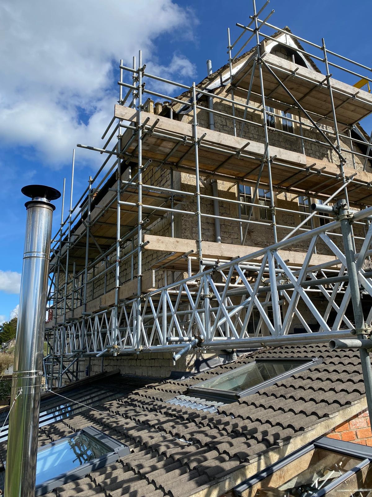 A house under construction or renovation is surrounded by scaffolding. The roof has skylights, and a shiny metal chimney is visible. The sky is mostly clear with some clouds.