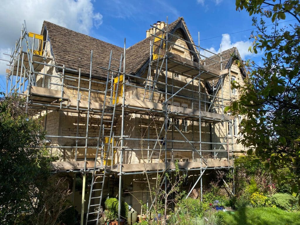 A large stone house with a steep, tiled roof is surrounded by scaffolding, indicating construction or renovation work. The house is set in a lush garden, and the sky is partly cloudy with sunlight shining through.