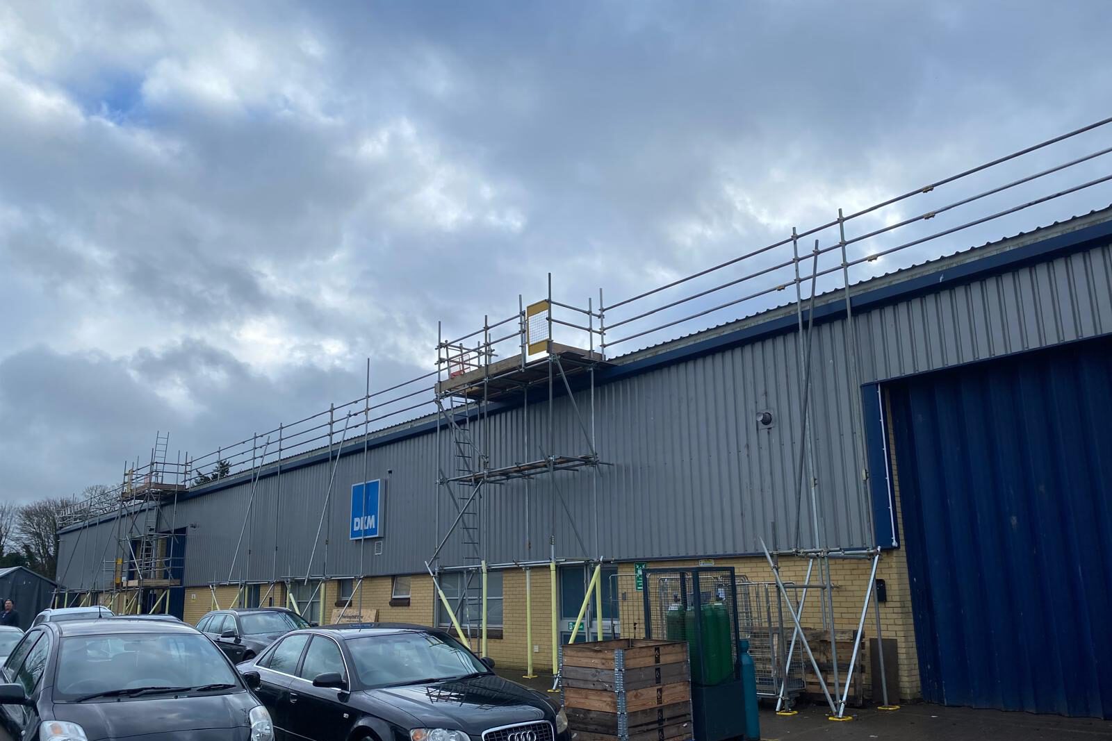 Scaffolding is set up along the side of a large industrial building with a blue and gray exterior. Several cars are parked in front, and the sky above is cloudy.