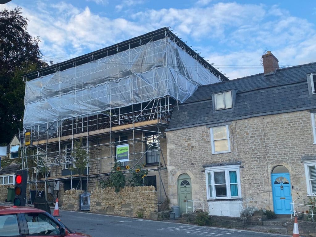 A stone building covered in scaffolding and white sheeting stands next to two older stone houses with blue doors. Sunflowers grow in front. A red traffic light and orange cones are visible on the street.