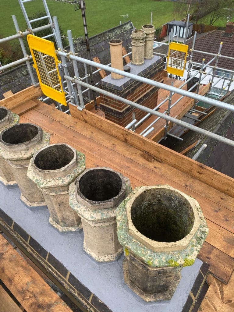 View of a rooftop under renovation with several old, weathered chimney pots in the foreground and scaffolding with safety barriers surrounding the area. Green fields and rooftops are visible in the background.