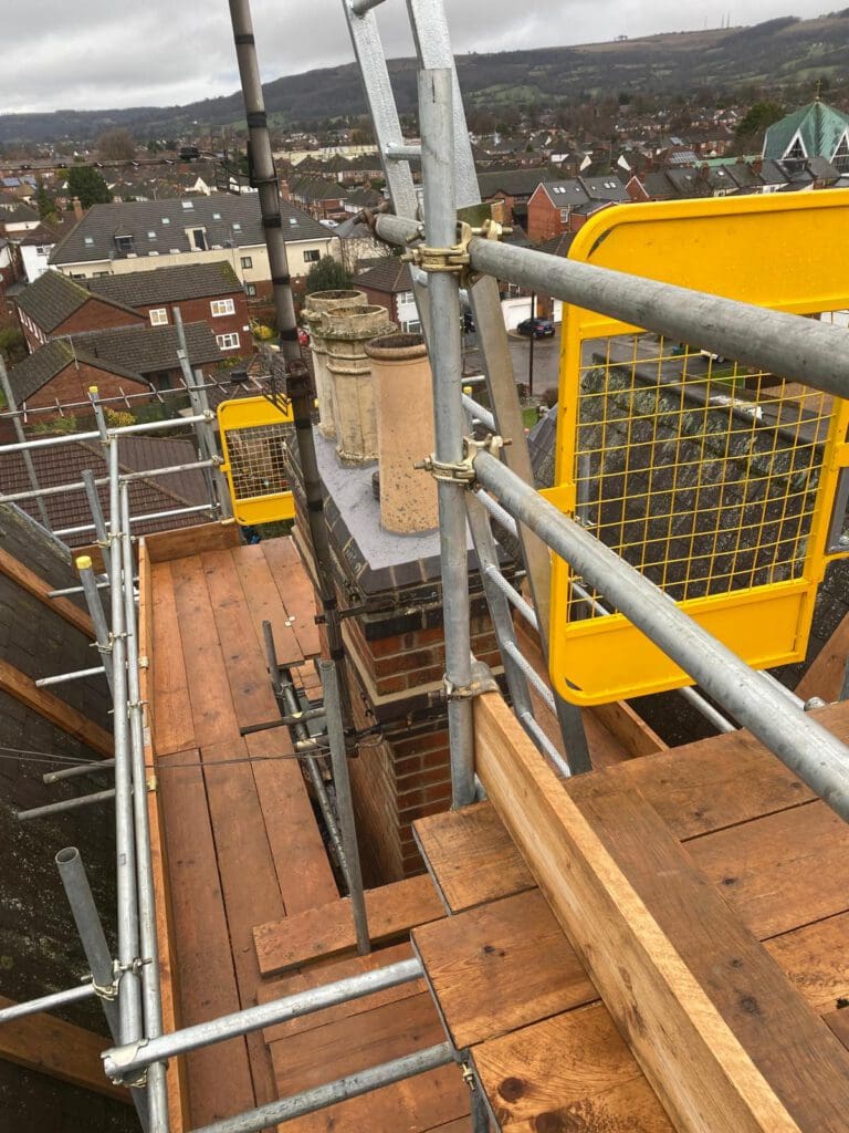 Scaffolding surrounds a rooftop chimney lined with pots, providing access for construction or maintenance work. Residential houses and roads are visible in the background under a cloudy sky.