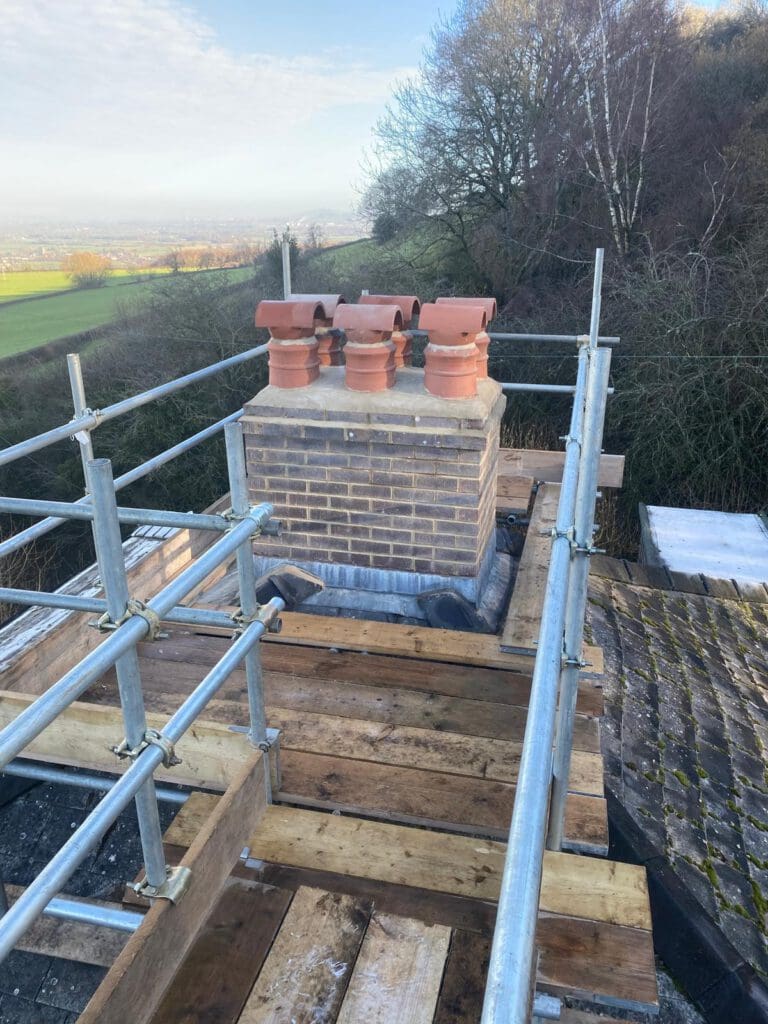 A brick chimney with three red clay chimney pots is surrounded by scaffolding and wooden planks on a rooftop, with trees and fields visible in the background under a partly cloudy sky.