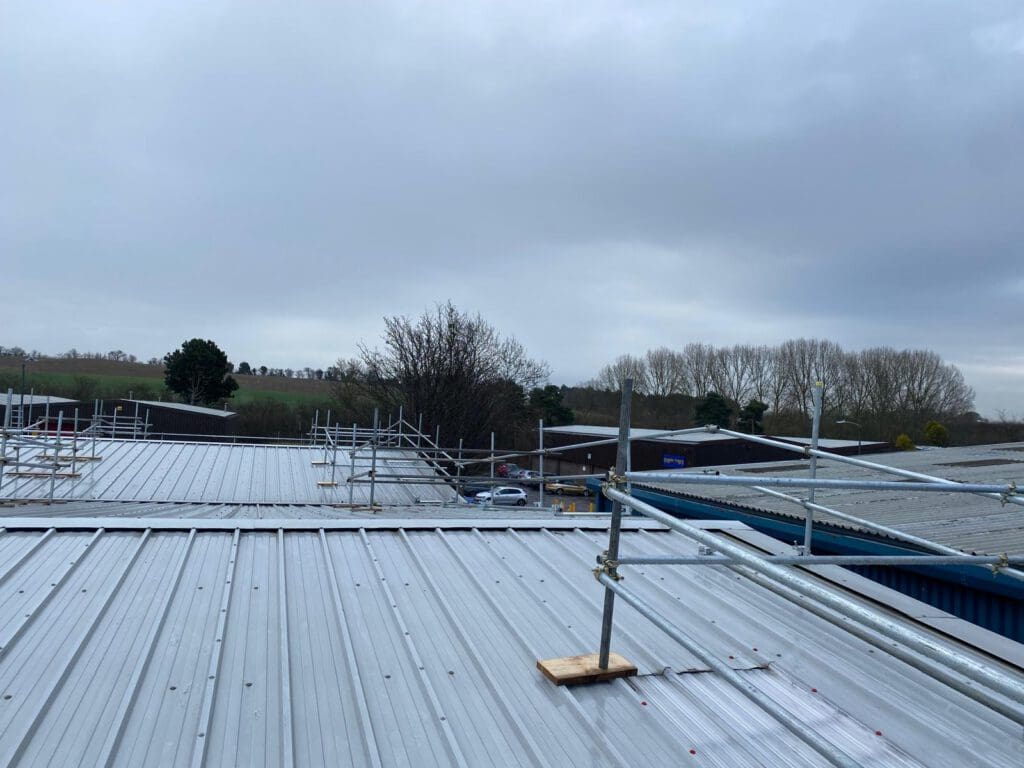 View from a rooftop with metal sheets and scaffolding; trees and fields are visible in the background under a cloudy sky. Some buildings and a parked car are also seen below.