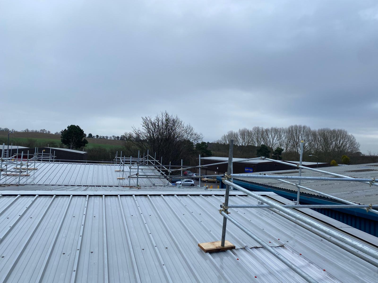 View from a rooftop with metal sheets and scaffolding; trees and fields are visible in the background under a cloudy sky. Some buildings and a parked car are also seen below.