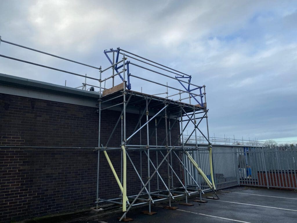 Scaffolding structure erected against the side of a brick building, with metal poles and platforms. The sky is cloudy, and a fenced area is visible in the background. The ground beneath is a paved surface.
