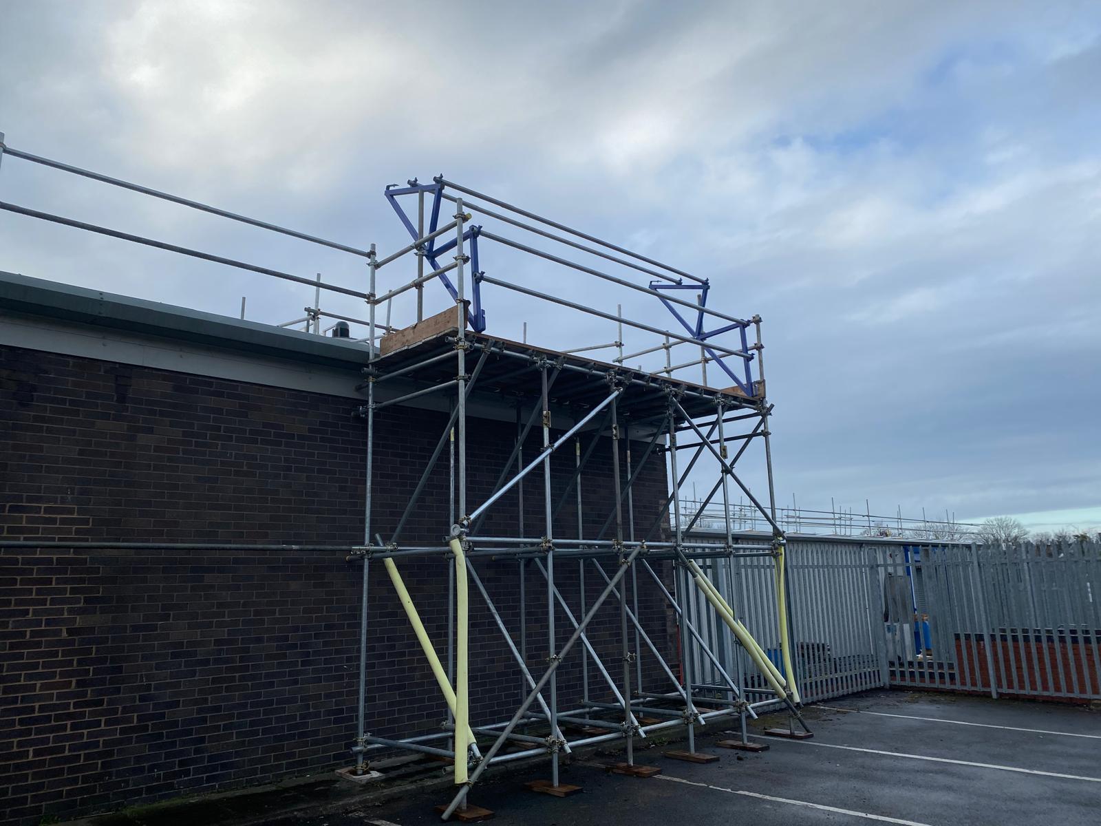 Scaffolding structure erected against the side of a brick building, with metal poles and platforms. The sky is cloudy, and a fenced area is visible in the background. The ground beneath is a paved surface.