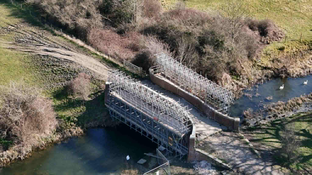 Aerial view of a footbridge under construction over a narrow river, surrounded by grass, trees, and muddy paths, with scaffolding covering most of the bridge.