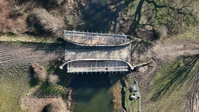 Aerial view of a partially demolished bridge over a river, with only the edges of the bridge remaining and gaps in the middle. Surrounding area is grassy with some trees and dirt paths.