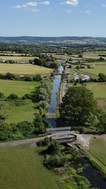 A scenic aerial view of a narrow river winding through green fields, with a small footbridge crossing over it and rolling hills visible in the background under a blue sky with scattered clouds.