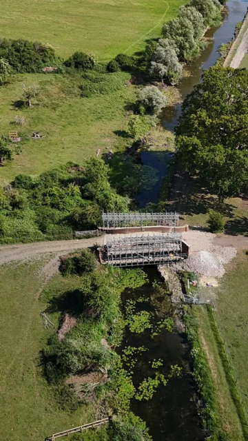 Aerial view of a small bridge over a narrow river, surrounded by green fields, trees, and patches of lily pads in the water. A path runs alongside the riverbank.