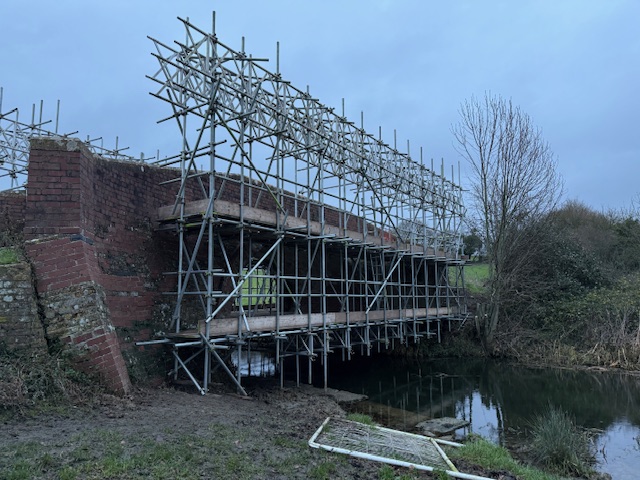 A brick bridge over a river is covered with extensive metal scaffolding, likely for repair or maintenance, with leafless trees and grassy fields in the background under a cloudy sky.