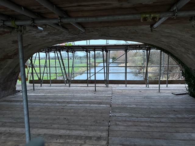 View from under a stone bridge with wooden planks and scaffolding, looking out toward a river and green fields in the distance.